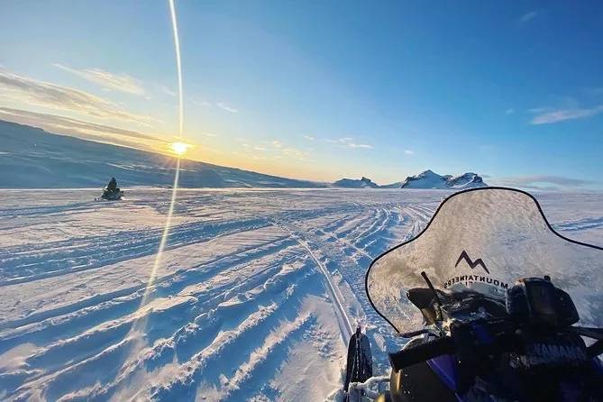 La vista dal sedile di una motoslitta: parabrezza, manubrio e tracce fresche che corrono verso un sole basso sul ghiaccio