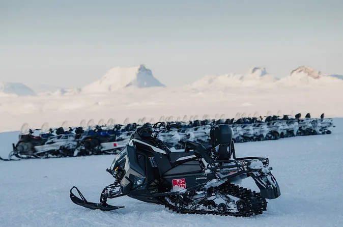 File di motoslitte parcheggiate in un campo base su un'ampia distesa di neve, con una montagna bassa all'orizzonte