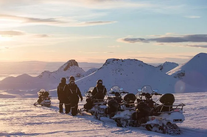 Diverse motoslitte parcheggiate sul ghiaccio al crepuscolo con i piloti in piedi accanto e cime innevate dietro