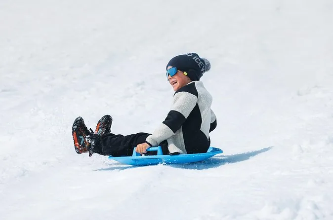 Ein Kind mit Mütze und Sonnenbrille rutscht auf einem blauen Plastikschlitten den Gletscherschnee hinunter