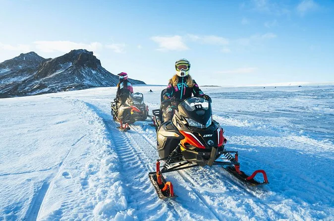 Zwei Fahrer auf einem roten Schneemobil auf einer präparierten Gletscherspur unterhalb eines dunklen felsigen Kamms