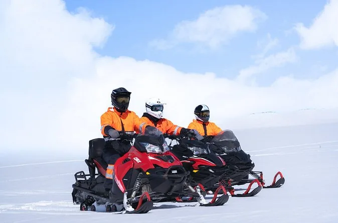 Drei rote Schneemobile nebeneinander auf dem Gletscher, jedes mit einem Fahrer und einem Beifahrer in orange-schwarzen Anzügen
