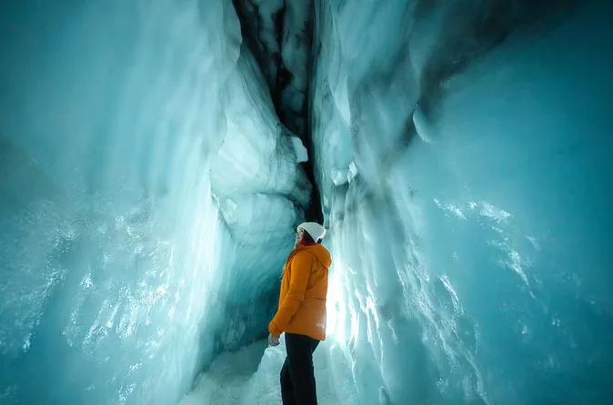 A visitor in an orange jacket standing in a narrow passage of deep blue glacier ice