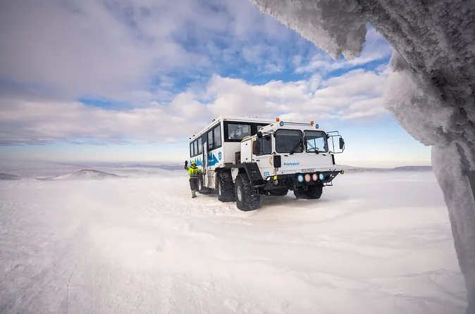 A white eight-wheeled glacier truck parked on the snow below an overhanging wall of glacier ice